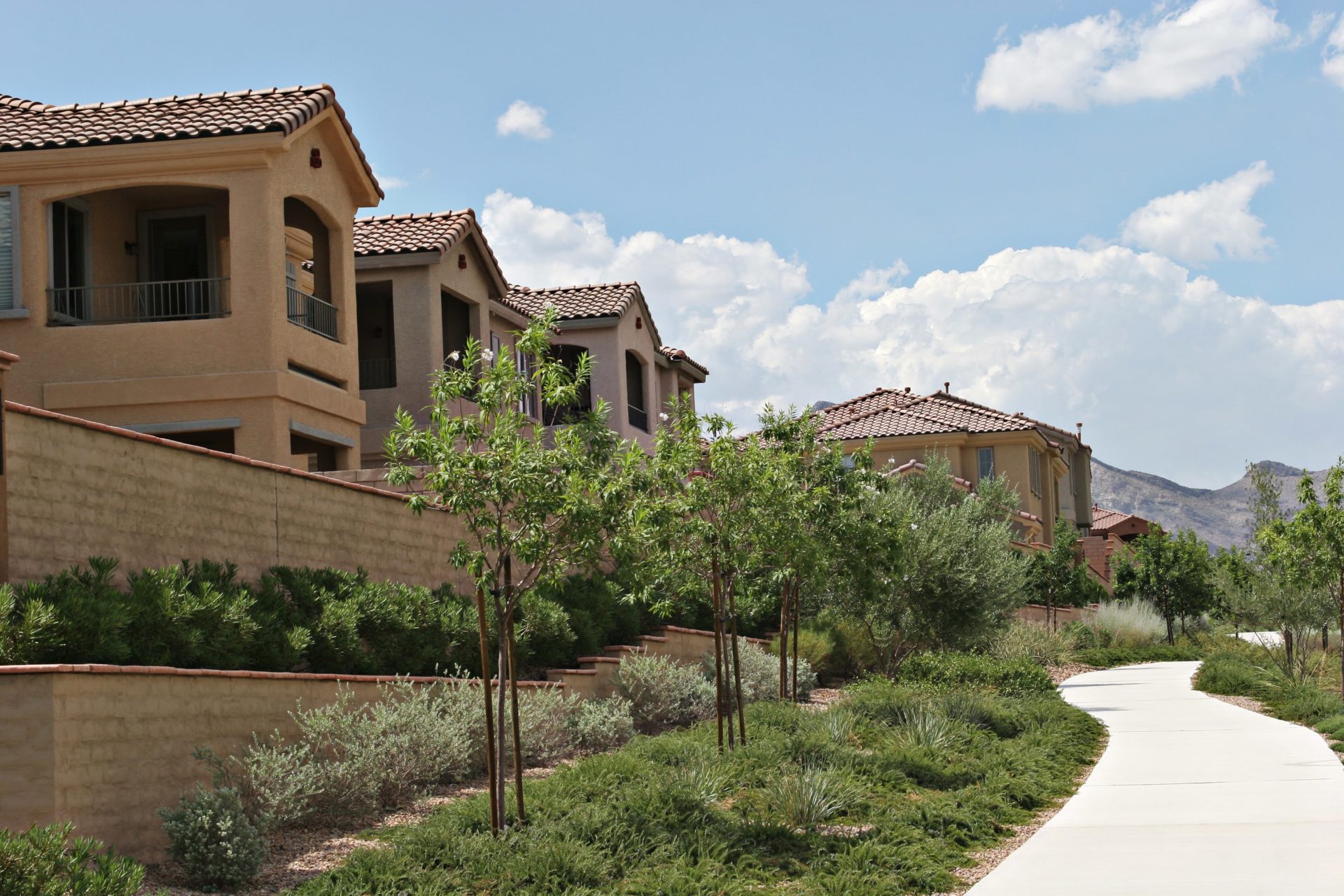 A tree-lined sidewalk path behind an apartment or condo complex that would benefit from New West Property Management services. The mission-style buildings are sided with beautiful brown stucco and terra-cotta tile roofs.