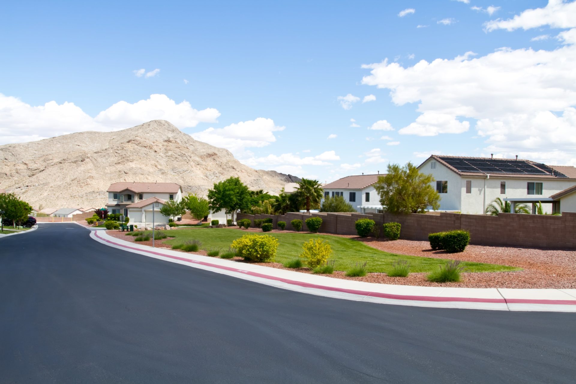 A street in a residential desert neighborhood. The three mission-style homes in view have white stucco siding and terra cotta tile roofs. The nearest house has solar panels. There is some green landscaping between the road and the houses